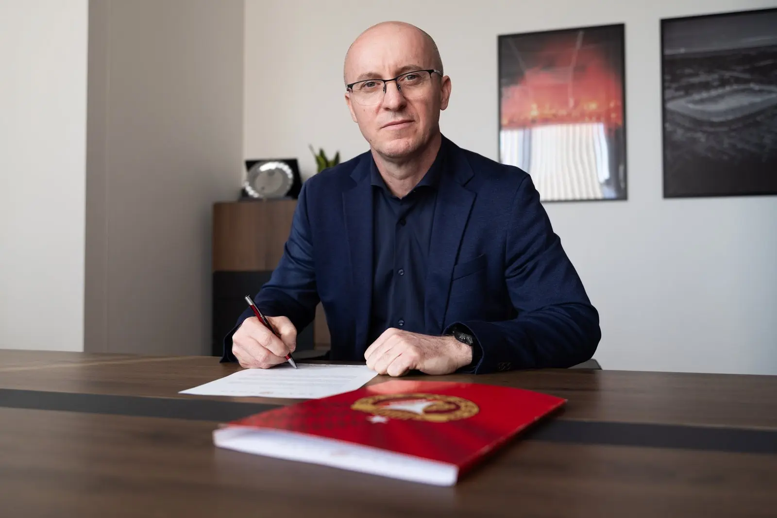 Aleksandar Vuković wearing a blue suit and glasses, sitting at a desk and signing a document
