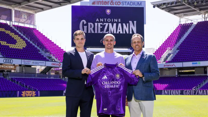 Antoine Griezmann posing with Orlando City officials holding a purple team jersey at Inter&Co Stadium
