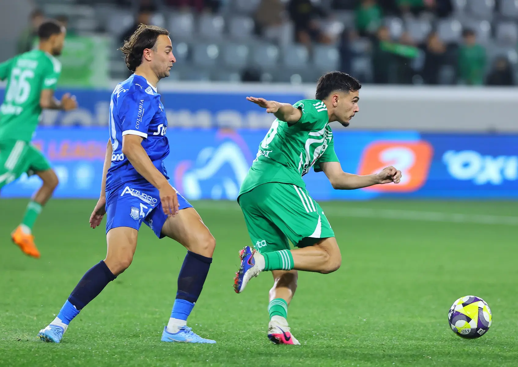 A soccer player in a green Aris Limassol kit dribbles the ball past a player in a blue Apollon Limassol kit.