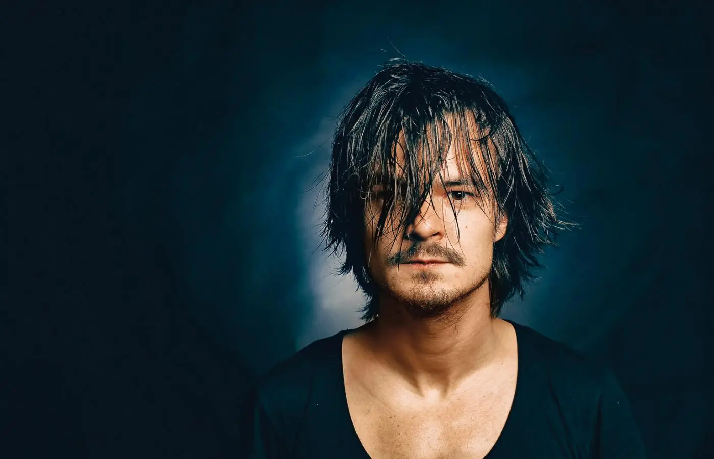 Portrait of actor Dawid Ogrodnik with dark, messy hair and a slight beard against a dark blue background