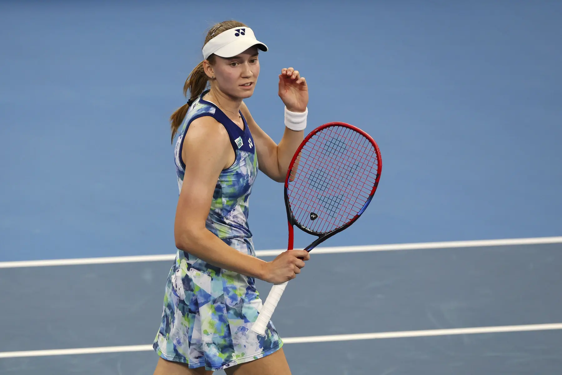Elena Rybakina on a blue tennis court holding her racket and wearing a white visor