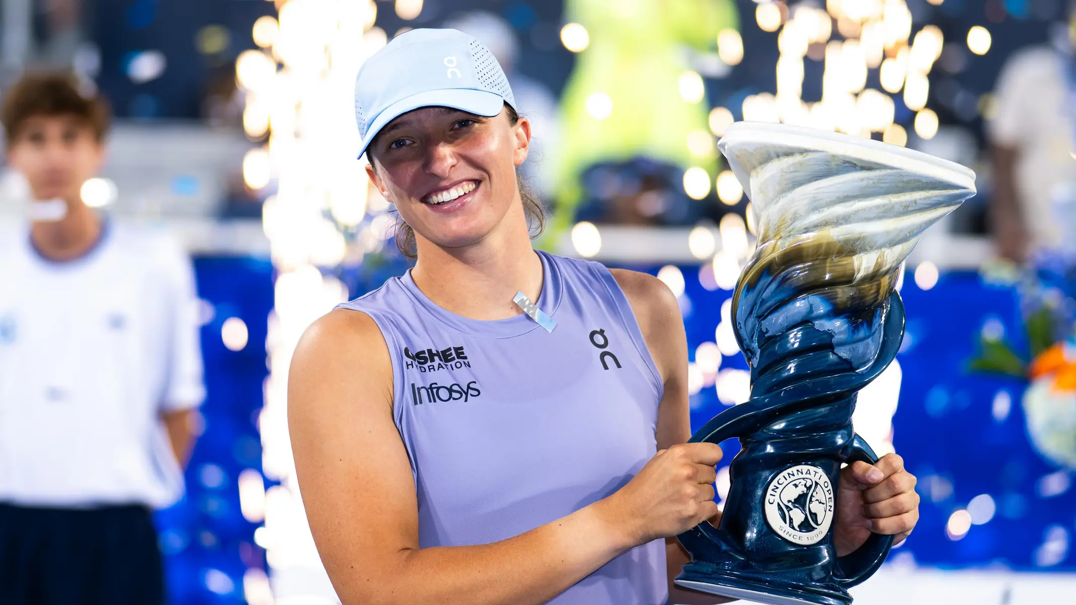 Iga Świątek smiling and holding the Cincinnati Open trophy while wearing a light blue tennis outfit and cap.
