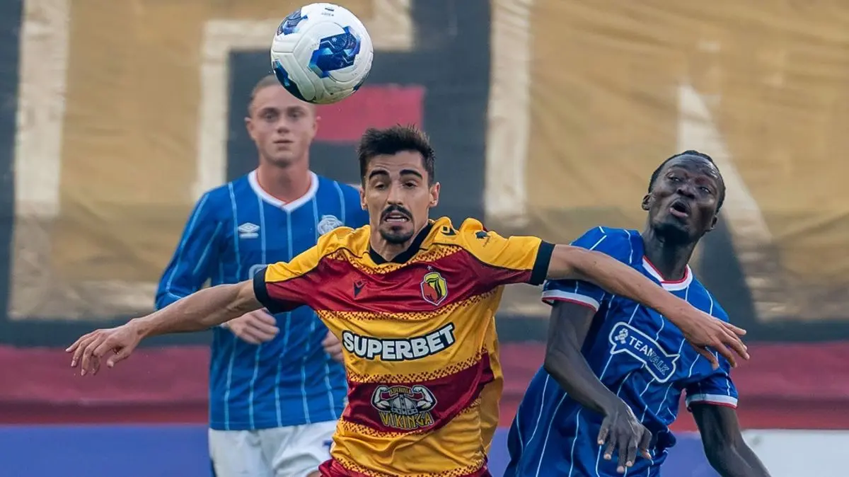 A Jagiellonia Białystok player in a yellow and red striped jersey contesting a header against two players in blue.