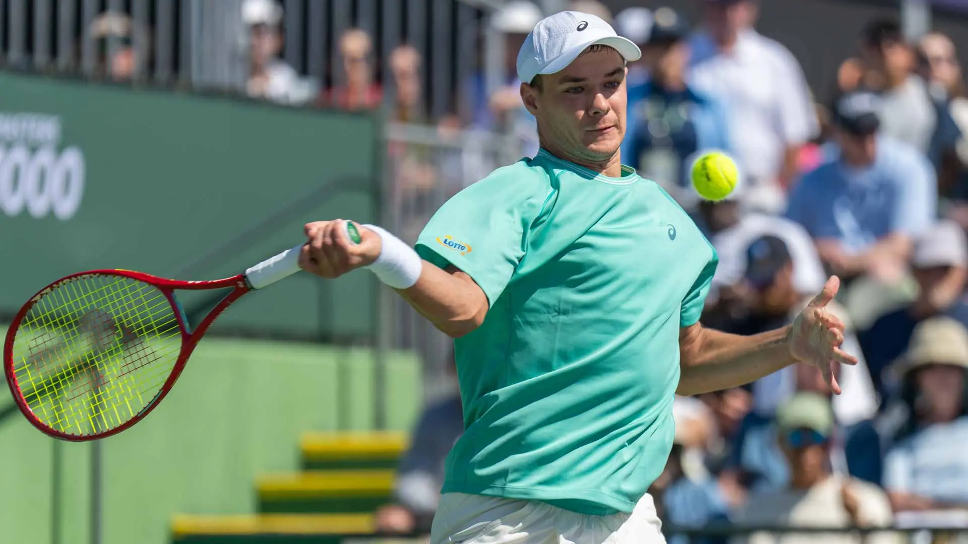 Kamil Majchrzak in a teal shirt and white cap hitting a tennis ball during a match.