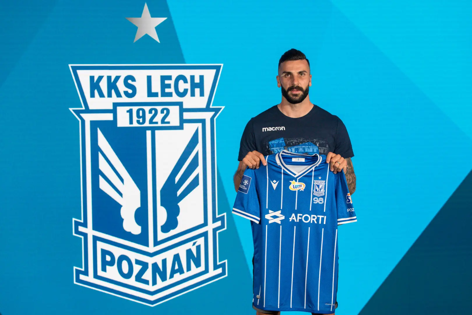 Mikael Ishak holding a blue and white striped Lech Poznań jersey in front of the club logo.