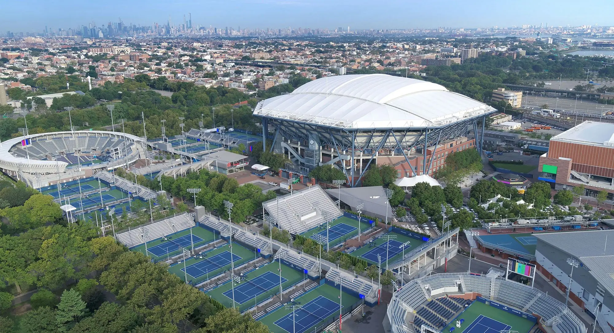 Aerial view of the US Open tennis complex including Arthur Ashe Stadium and multiple blue hard courts.