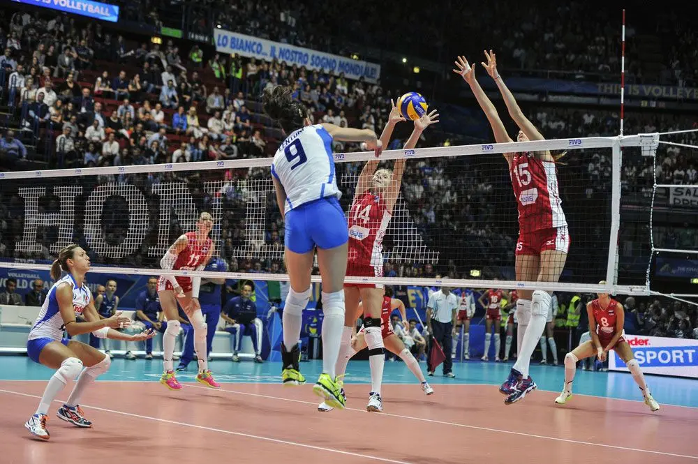 Female volleyball player in blue jersey spiking the ball over a net against blockers in red jerseys