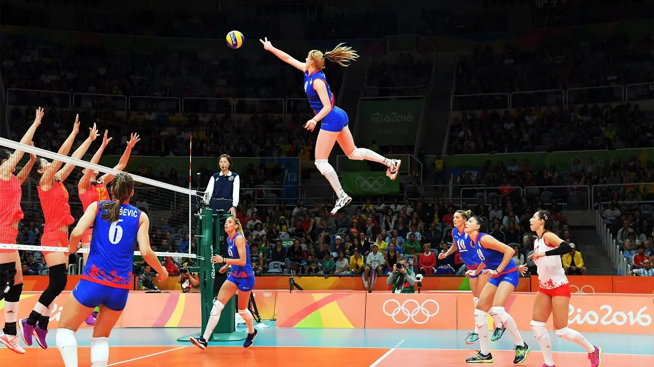 A female volleyball player in a blue uniform jumps high to spike the ball over the net during a match.