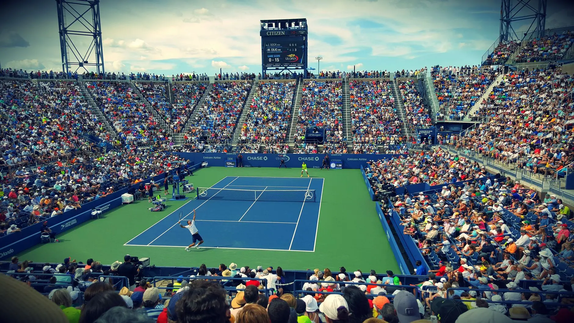 Wide shot of a tennis match in progress at a large outdoor stadium with a full crowd.
